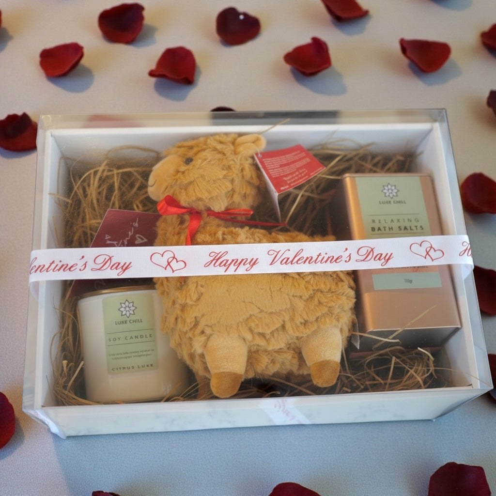 Valentine's Day gift set with a candle, plush toy, and bath salts on a white surface with red rose petals.
